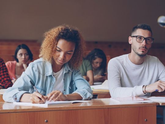 Uczniowie siedzący w sali wykładowej, skupieni na rozwiązywaniu zadań i zapisywaniu odpowiedzi podczas zajęć, z prowadzącym widocznym przy pierwszej ławce. Ujęcie podkreśla koncentrację i pracę w grupie, charakterystyczne dla skutecznego przygotowania do matury w Szczecinie – więcej informacji dostę